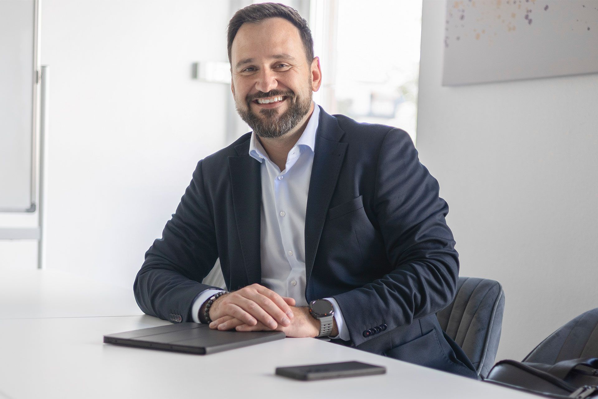 Oliver Bajunovic sitzt lächelnd am Besprechungstisch, vor ihm Tablet und Smartphone